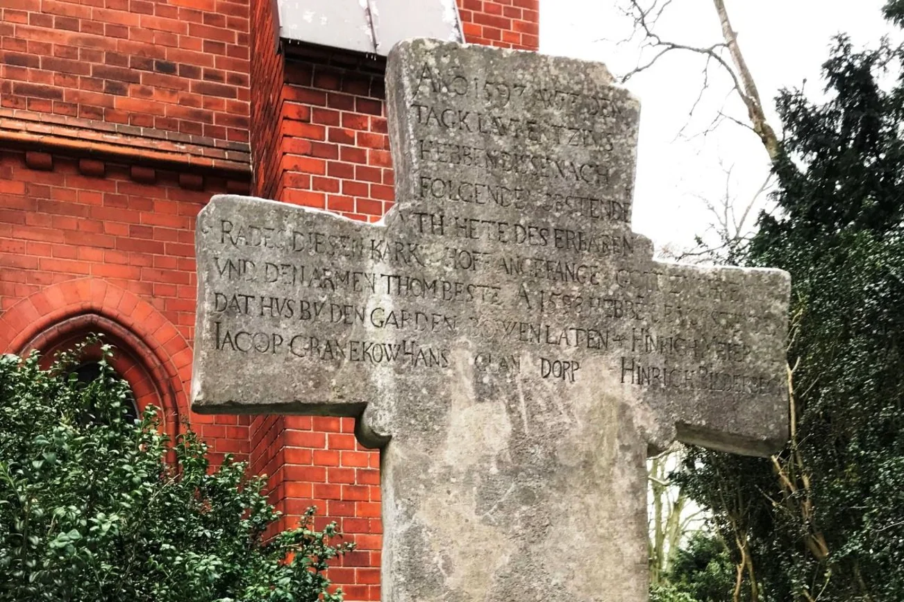 Ein Steinkreuz mit verwitterter Schrift, im Hintergrund eine rote Backsteinmauer.