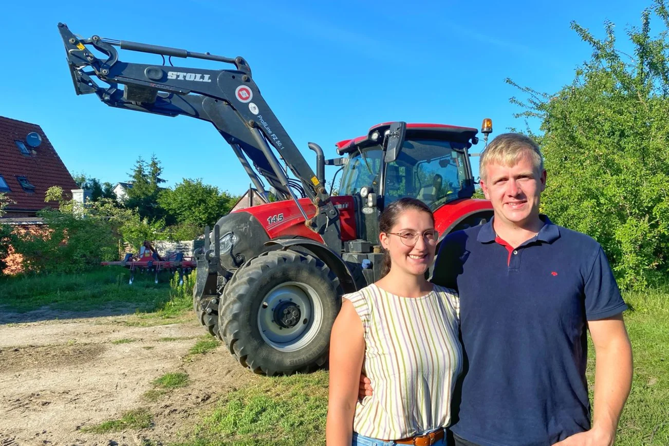 Zwei junge Menschen stehen lächelnd vor einem Trecker. 