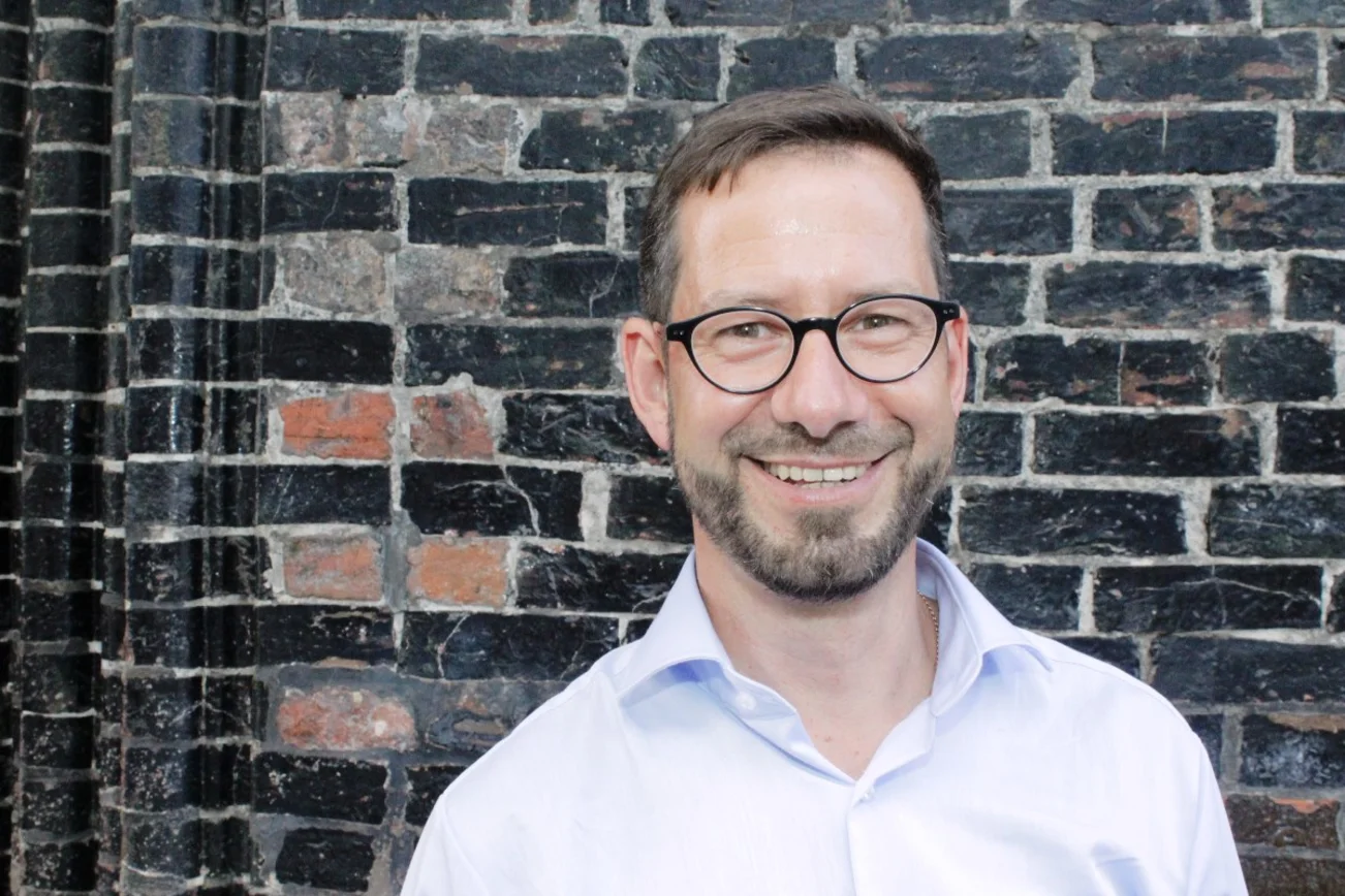 Das Portrait eines Mannes mit kurzen, dunklen Haaren und Brille.