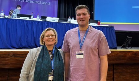 Eine Frau und ein Mann stehen in einem Sitzungssaal.