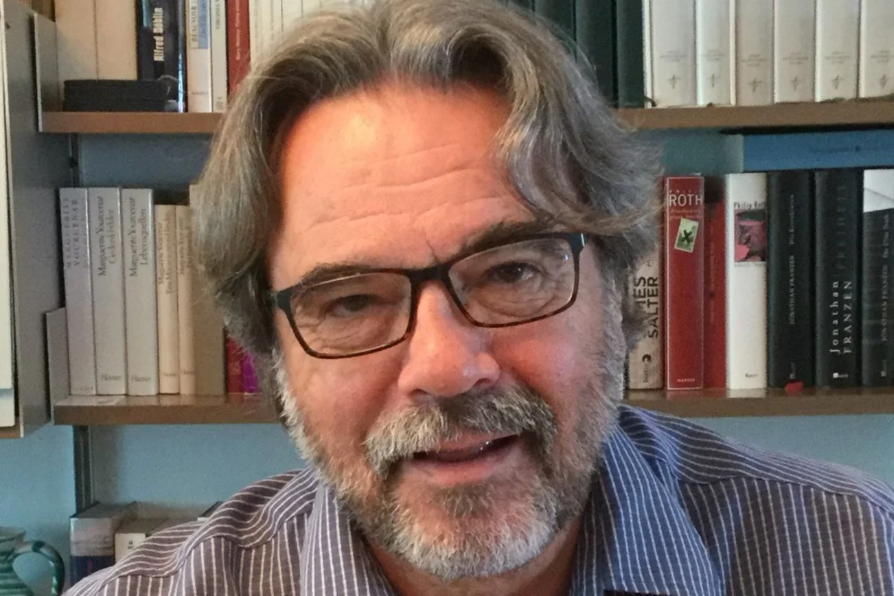 Das Portrait eines Mannes mit Brille. Im Hintergrund ein Regal mit Büchern.