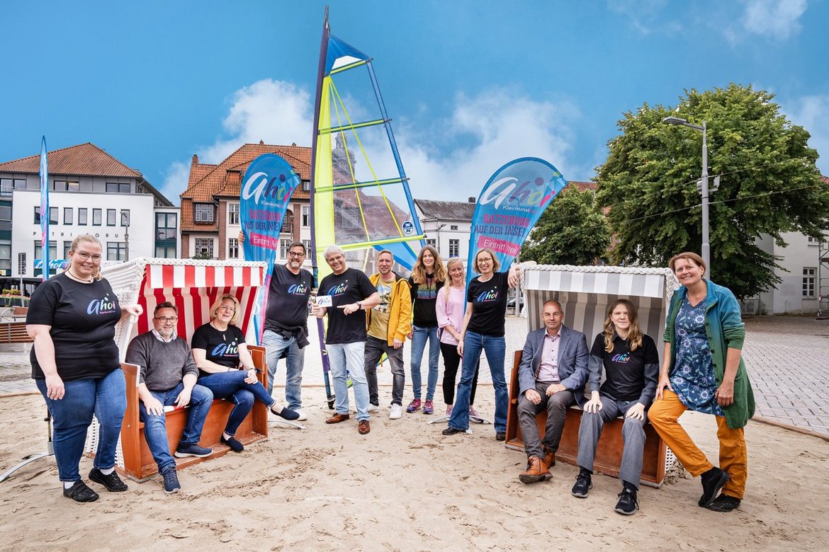 Zwei Strandkörbe links und rechts auf denen jeweils zwei Personen sitzen. Links, rechts und zwischen ihnen stehen lächelnde Menschen. Sie stehen auf Strandsand, im Hintergrund eine Häuser und davor ein Segel. Es ist blauer Himmel mit ein paar wenigen weißen Wolken