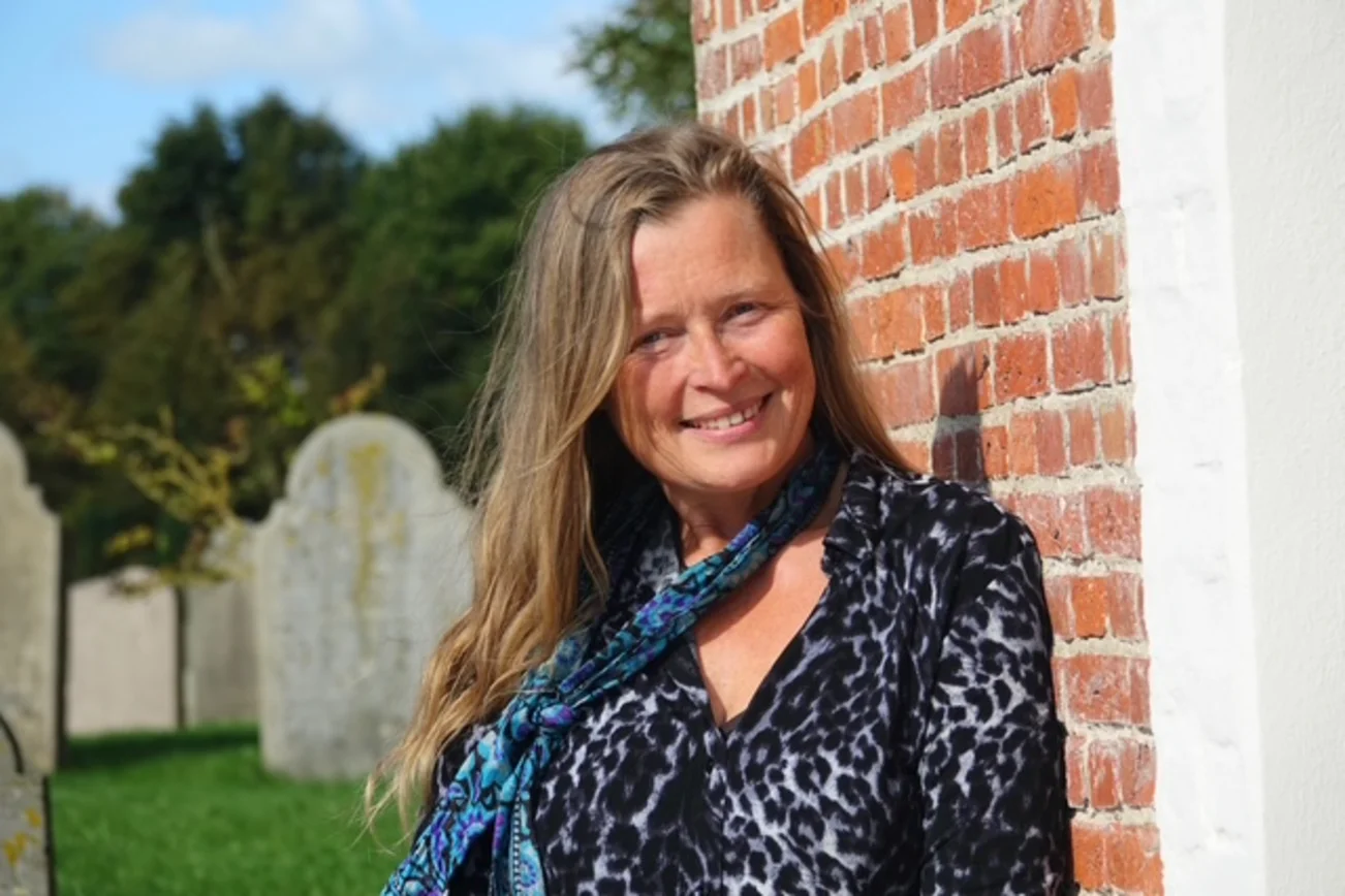 Frau lächelt an eine Backsteinmauer gelehnt in die Kamera.