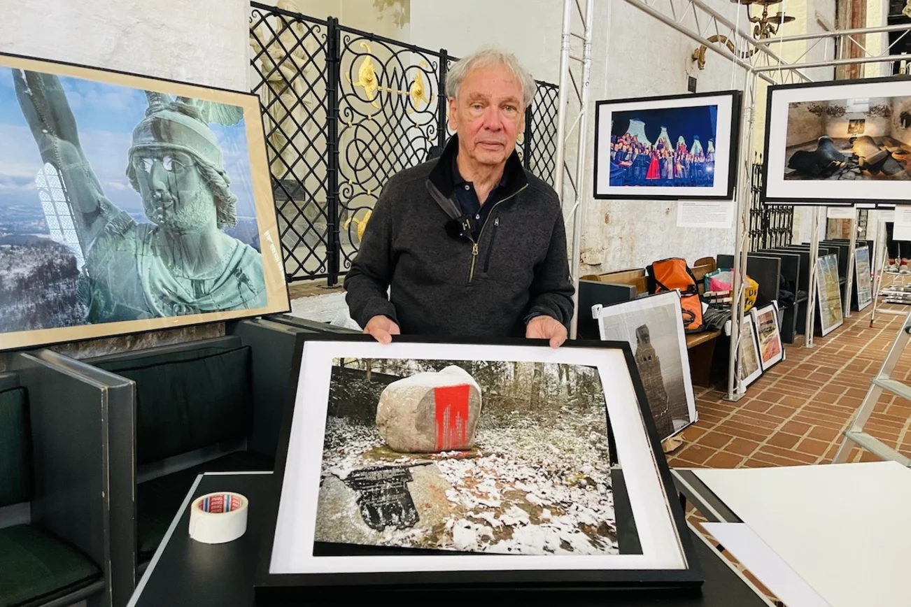 Fotograf Harald Schmitt steht vor seiner Ausstellung mit vielen Fotos von Orten des Gedenkens und hält ein Bild hoch.