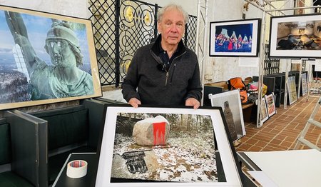Fotograf Harald Schmitt steht vor seiner Ausstellung mit vielen Fotos von Orten des Gedenkens und hält ein Bild hoch.