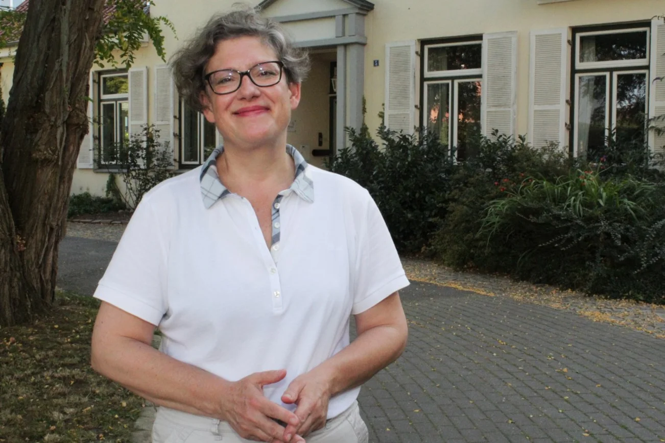 Eine Frau mit Brille. Sie trägt ein weißes Polo-Shirt. Im Hintergrund ein Haus.