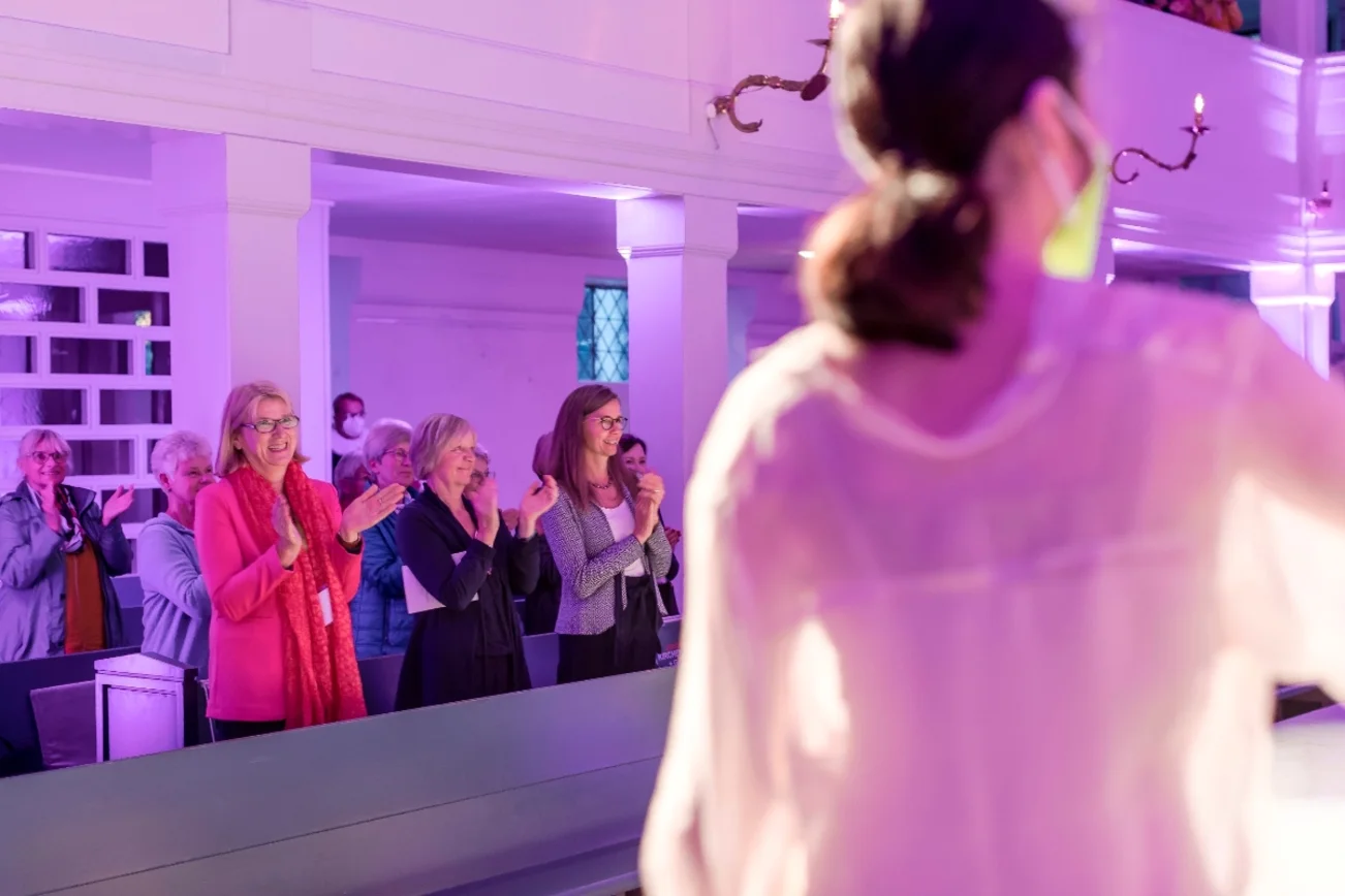 Vorn rechts im Bild unscharf ein Teil des Rückens einer Balletttänzerin. Im Hintergrund klatschen die Besucher:innen Beifall.