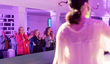 Vorn rechts im Bild unscharf ein Teil des Rückens einer Balletttänzerin. Im Hintergrund klatschen die Besucher:innen Beifall.