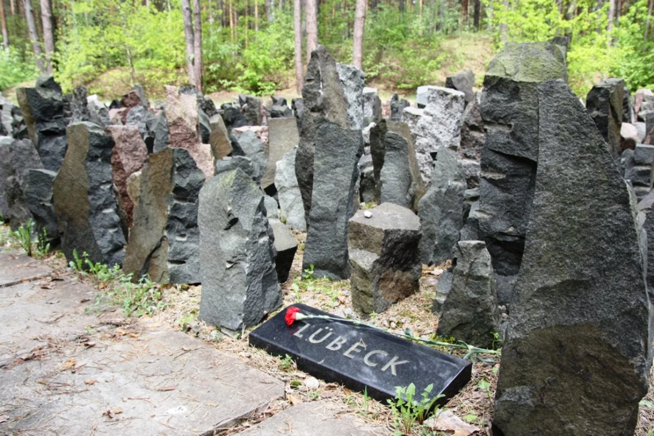 graue Steine stehe aufrecht lose nebeneinander. Eine Platte mit der Aufschrift Lübeck. Darauf liege eine rote Blume. Im Hintergrund Wald.