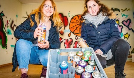 Zwei junge Frauen lächeln in die Kamera und halten Farbdosen in den Händen. 