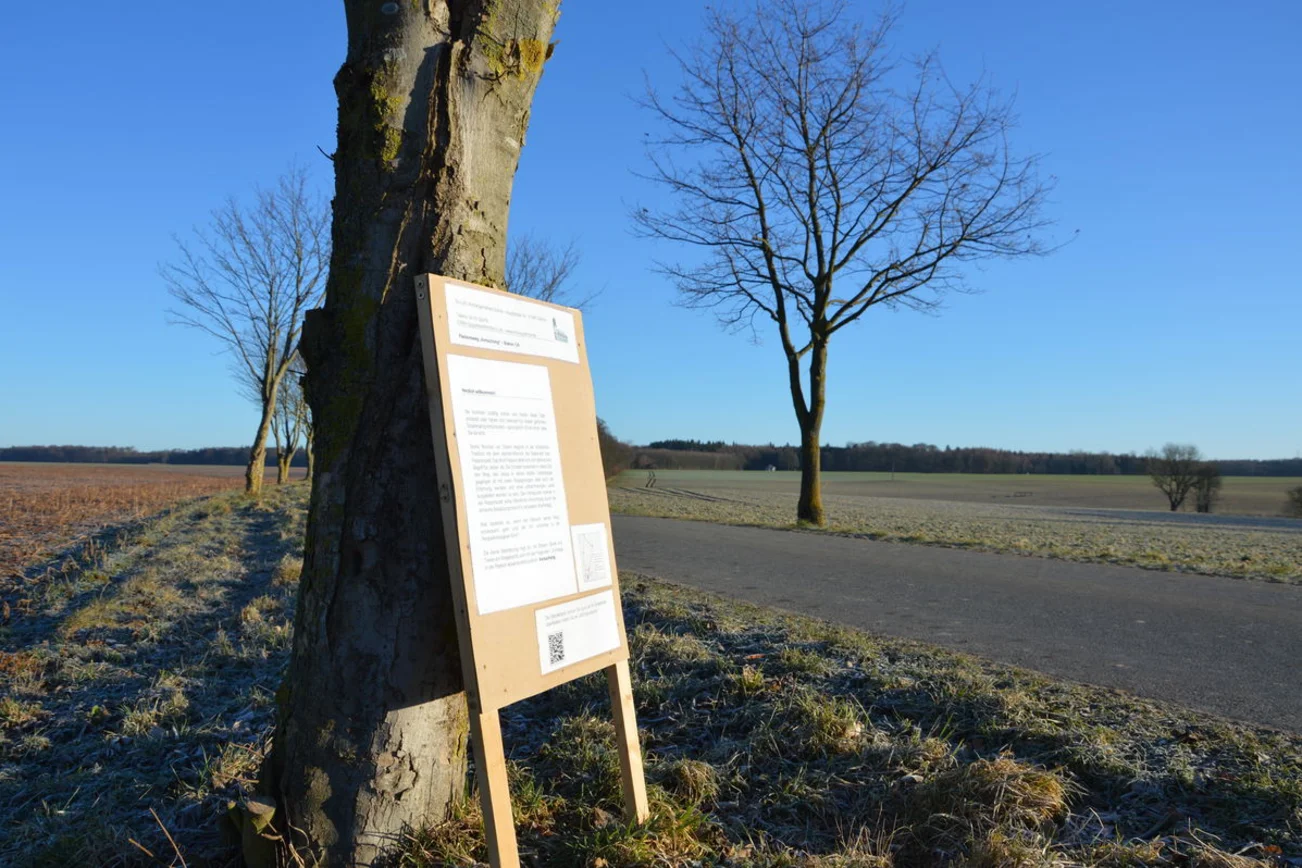 Man sieht eine Straße in der Landschaft. Im Vordergrund lehnt ein Aufsteller mit Texten an einem Baum