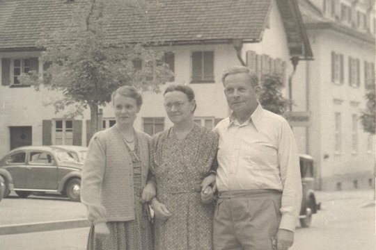 Ein historisches Foto. Drei Menschen sind zu sehen. Sie stehen eingeharkt nebeneinander.