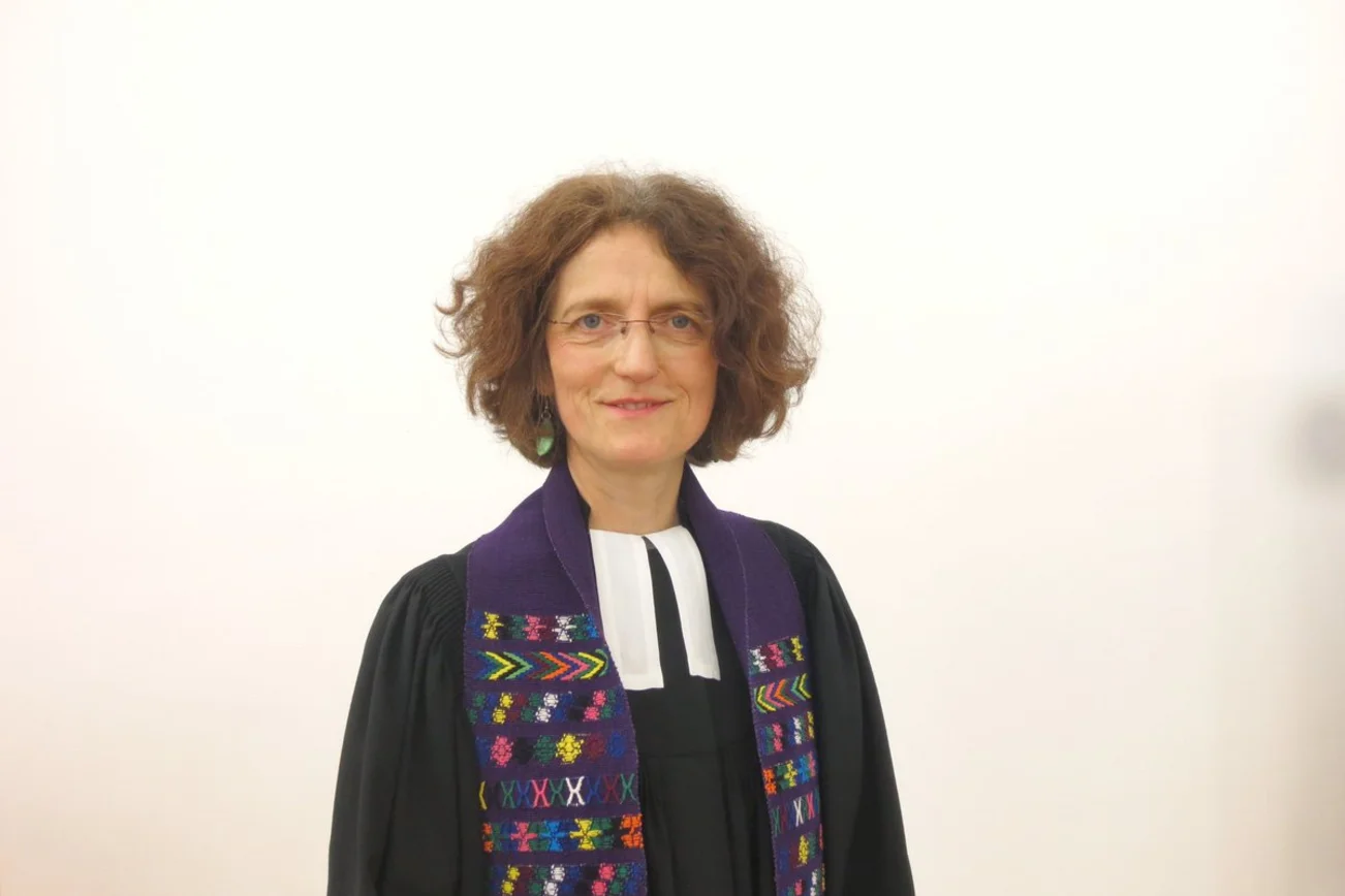 Eine Pastorin steht sanft lächelnd in einem Talar vor einer weißen Wand. 