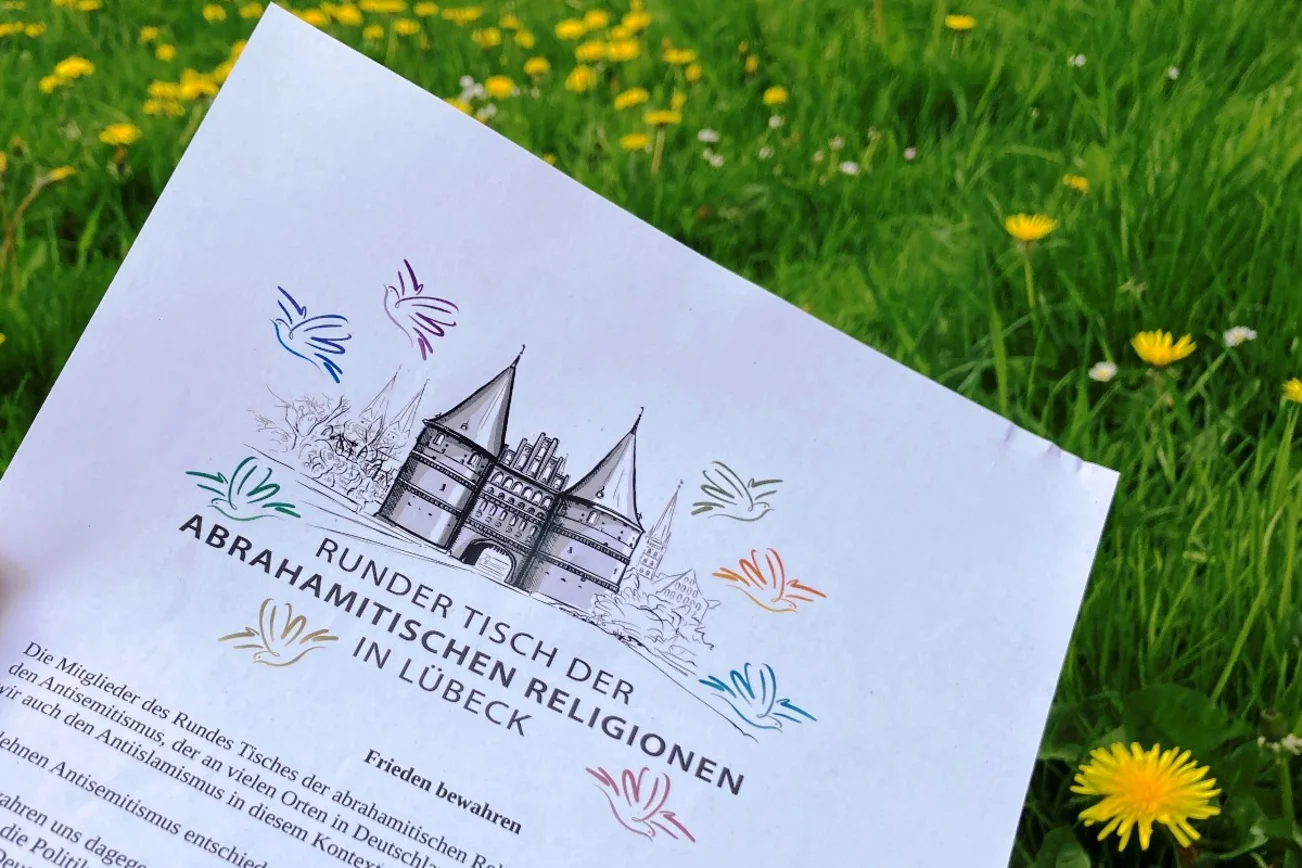 Ein Blatt Papier mit dem Logo des runden Tisches der abrahamitischen Religionen in Lübeck. Es zeigt eine Zeichnung des Holstentores umgeben von Tauben.