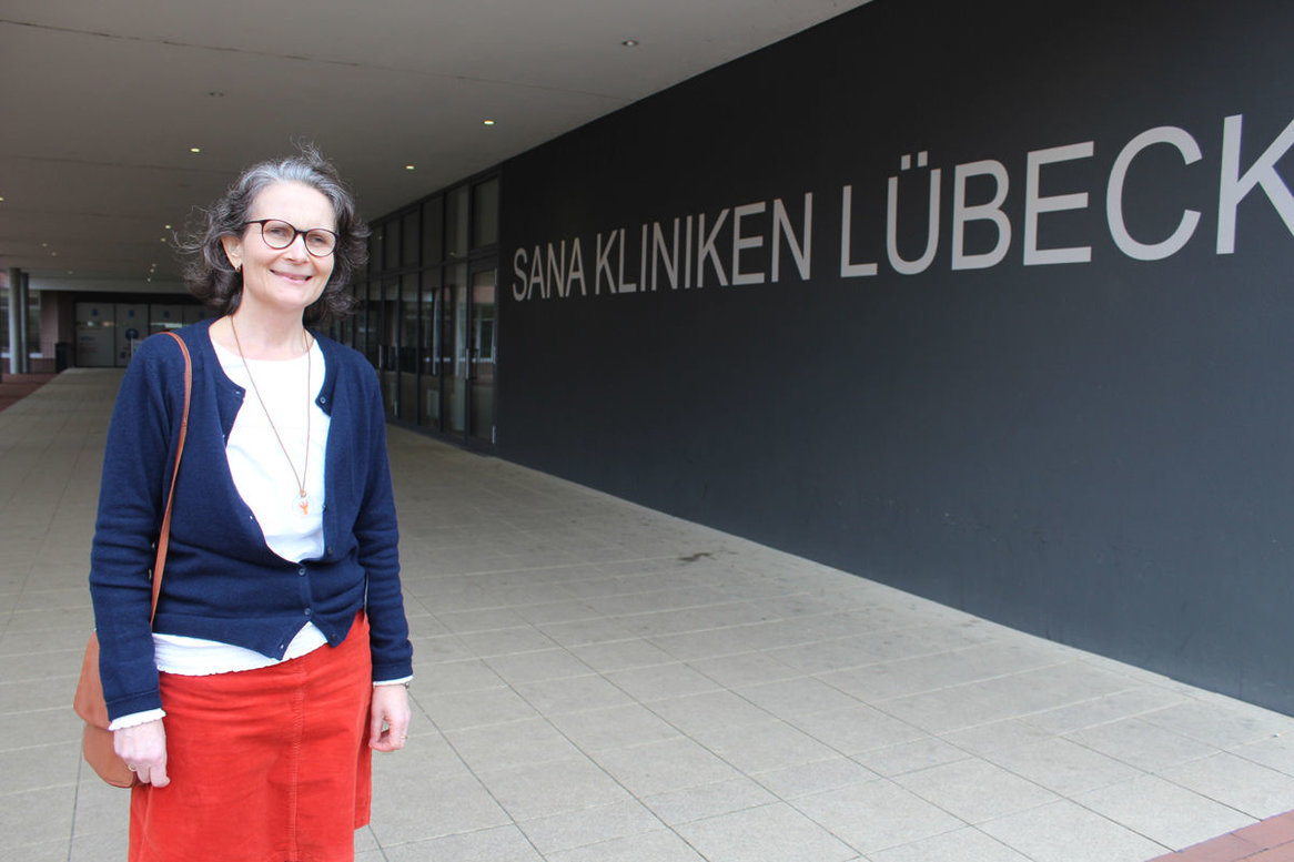 Links steht Frau Grossmann mit weißem Oberteil, blauer Stickjacke und rotem Rock, rechts ist der Schriftzug Sana Kliniken Lübeck in Großbuchstaben und weißer Schrift auf dunkelgrauer Wand zu sehen