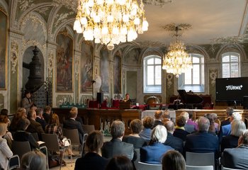 Eine Reihe von Menschen sitzt in einem barocken festlichem Saal, zwei Kronleuchter hängen von der Decke - Copyright: Oliver Beck 
