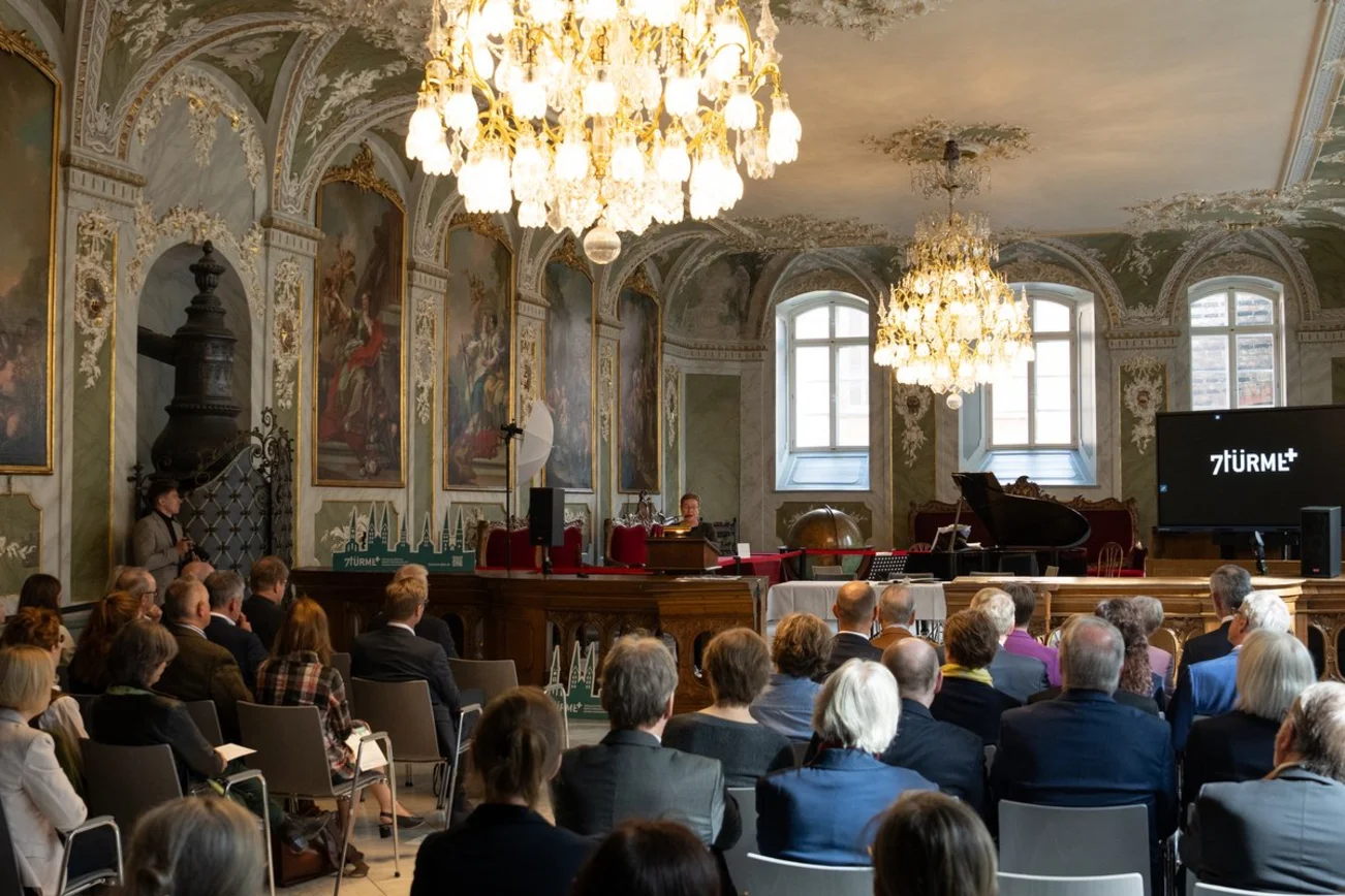 Eine Reihe von Menschen sitzt in einem barocken festlichem Saal, zwei Kronleuchter hängen von der Decke