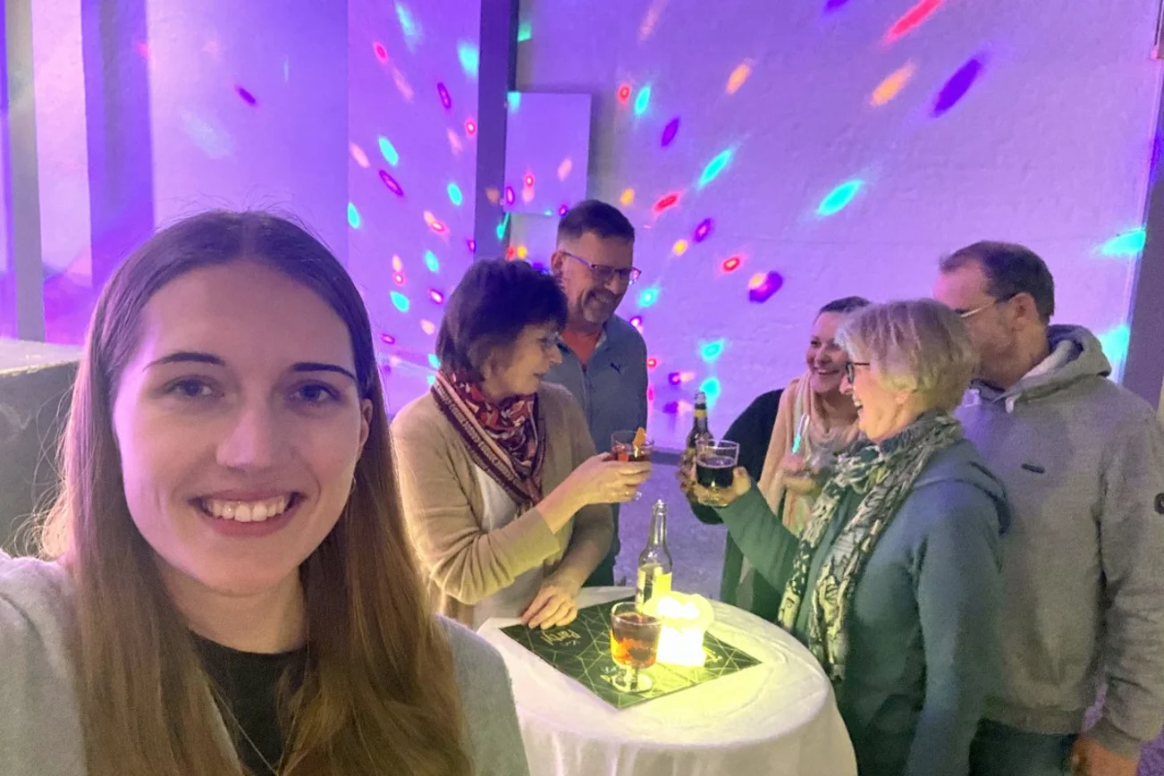 Junge Frau lächelt in Kamera, im Hintergrund steht eine Gruppe um einen Tisch herum.