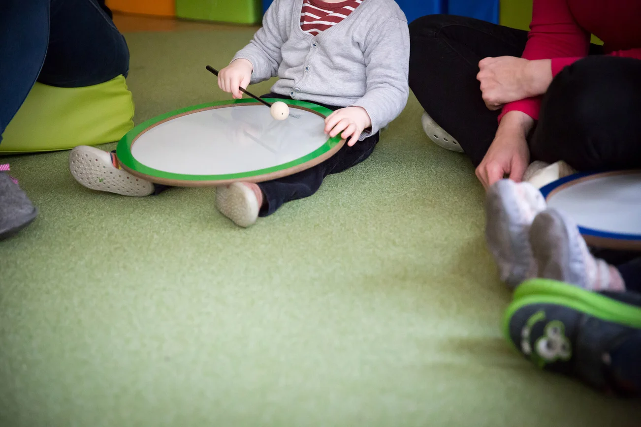 Kleinkind spielt mit einem Schlegel auf einer Knietrommel.