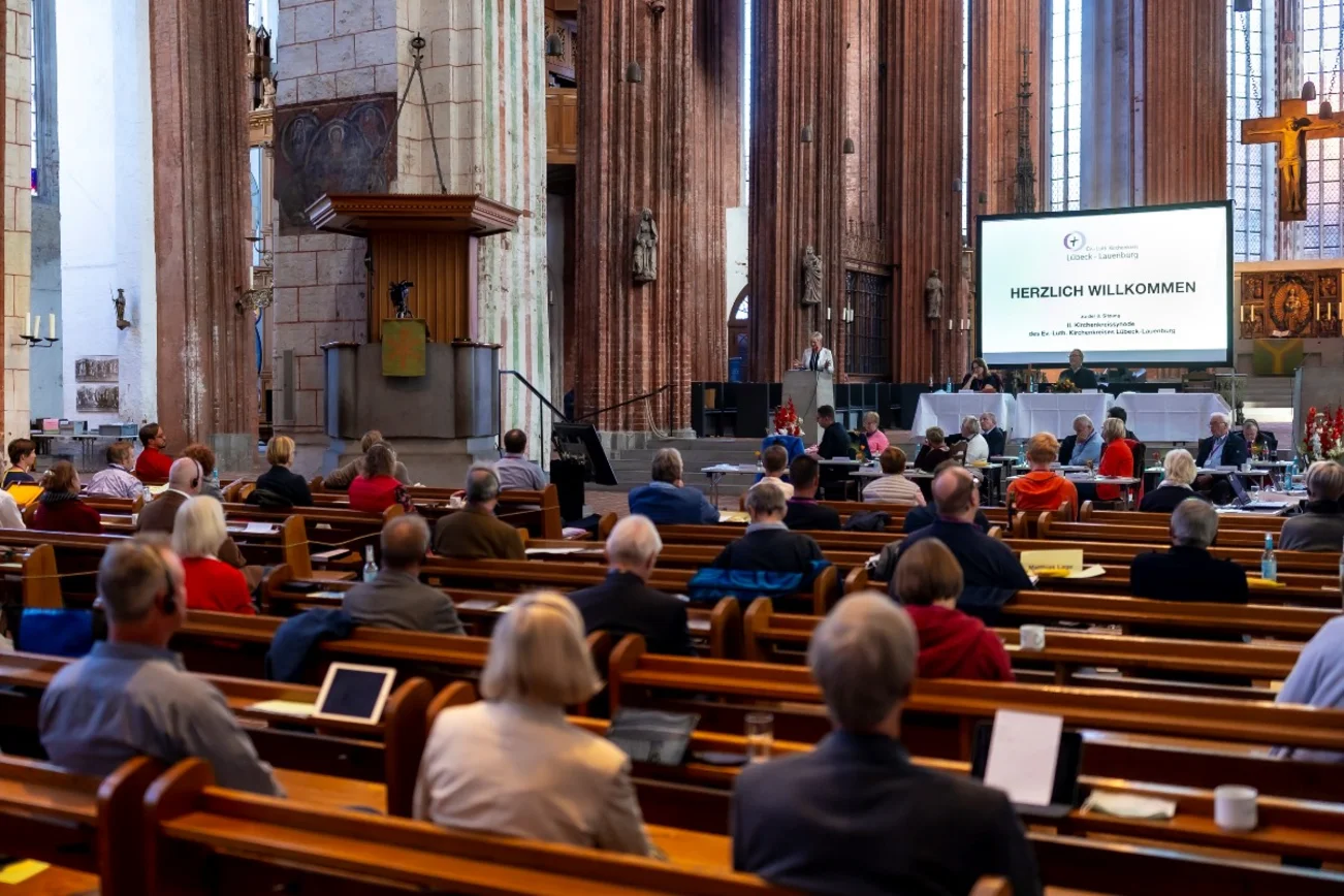 Ein Kirchraum, Menschen sitzen in Bänken auf Abstand. Im Vordergrund eine Leinwand.