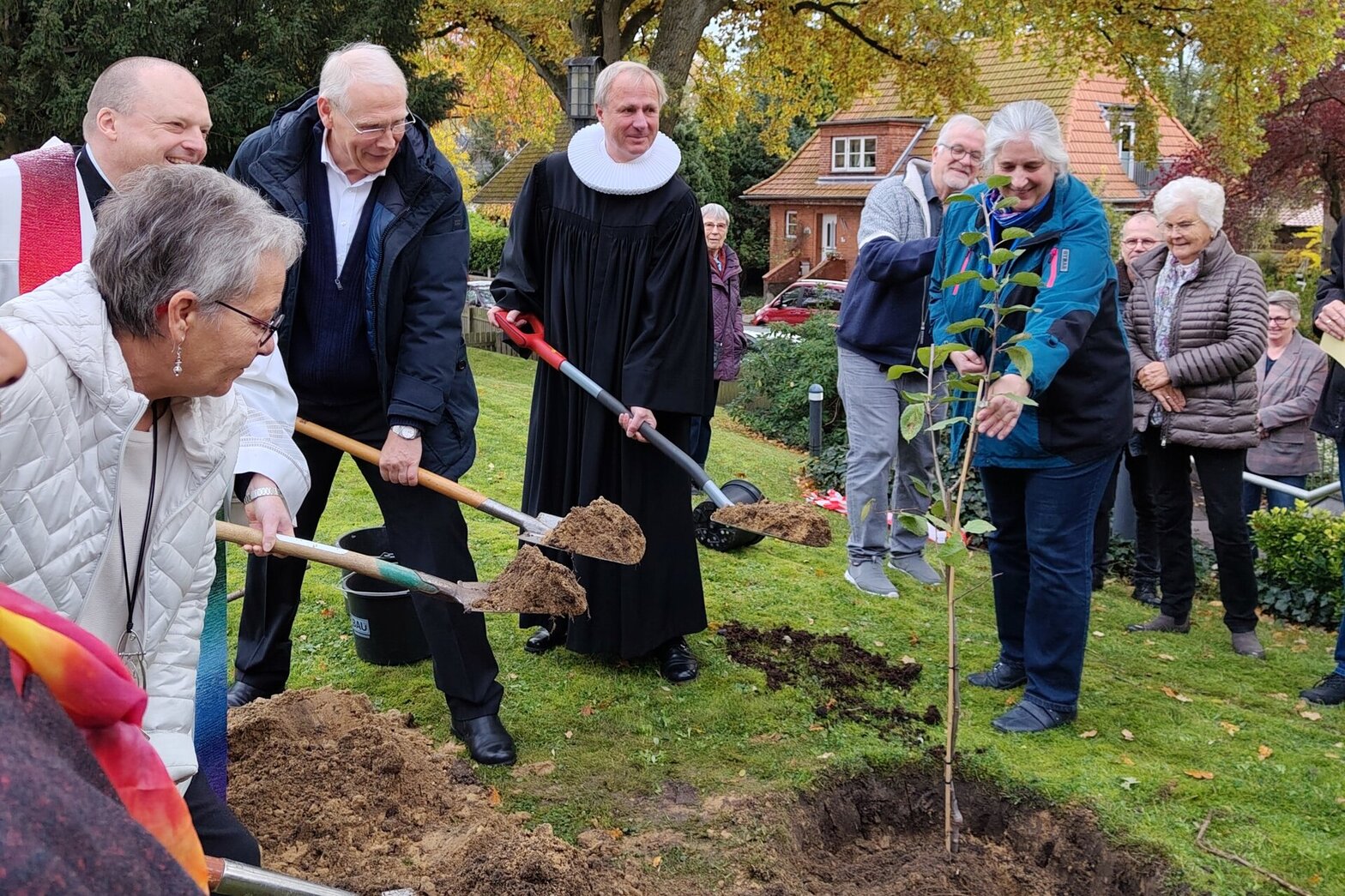 Viele Menschen mit Schaufeln in der Hand vor einem gegrabenen Loch mit einem kleinen Apfelbaum darin.
