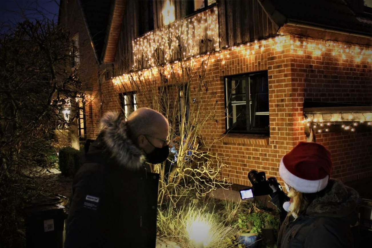 Vor einem weihnachtlich geschmückten Haus stehen zwei Menschen und blicken auf das Display einer Kamera