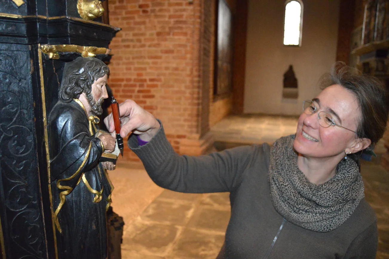 Eine Frau reicht einer kleinen Figur in einer Kirche einen Kugelschreiber.
