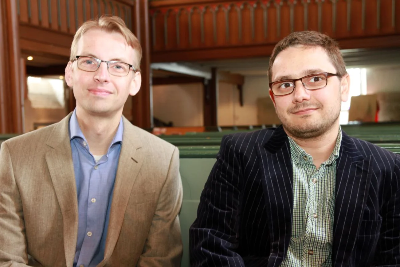Zwei Männer sitzen auf einer Kirchenbank und blicken freundlich in die Kamera. Im Hintergrund der Raum einer Kirche.