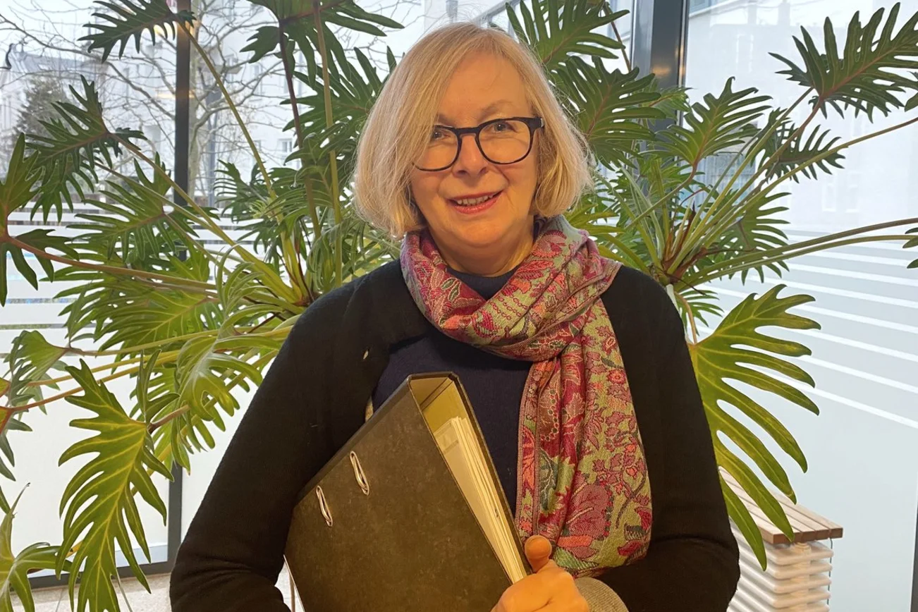 Eine Frau steht vor einer Pflanze und trägt einen Aktenordner unter dem Arm.