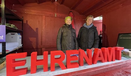 Zwei Frauen stehen in einer Holzhütte, im Vordergrund stehen die roten Buchstaben EHRENAMT