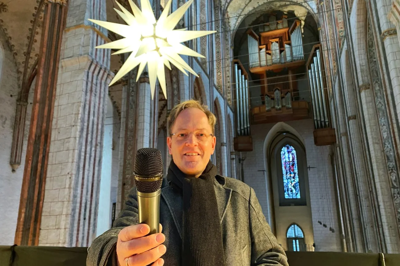 Mann steht in einer Kirche vor einem Leuchtstern und hält lächelnd ein Mikrofon in die Kamera