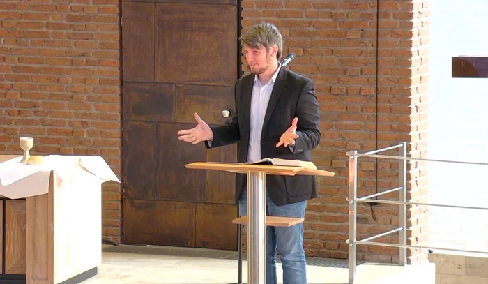 Ein Mann steht vor einem Pult und hält die Arme vor sich geöffnet und schaut nach vorne