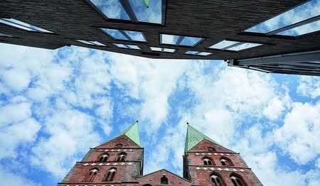 Die Türme von St. Marien und ein Haus der Innenstadt ragen in den wolkenverhangenen Himmel, welcher an vielen Stellen blauen Himmel erkennen lässt. 