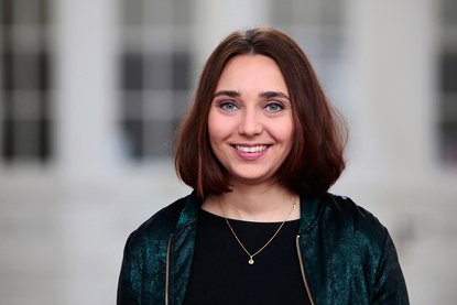 Eine Grau mit schulterlangen braunen Haaren und dunkler Kleidung steht vor einem weißen Hintergrund, der etwas unscharf zu erkennen ist und lächelt in die Kamera - Copyright: Anne Oschatz