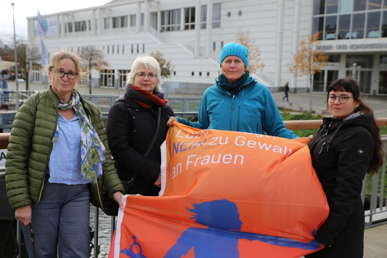 Vier Frauen stehen mit einer orangefarbenen Fahne vor der Lübecker Musik- und Kongresshalle.