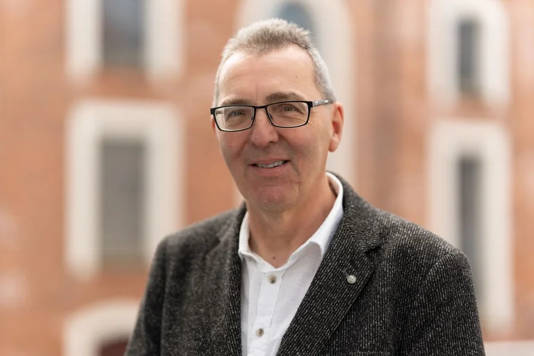 Ein Mann mit einer Brille und kurzen grauen Stoppelhaaren. Er grägt ein weißes Hemd und ein dunkles Oberteil darüber