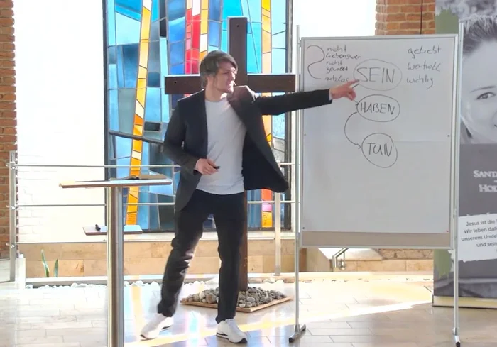 Ein Mann steht vor einem Holzkreuz und einer Tafel und zeigt mit dem Finger auf diese