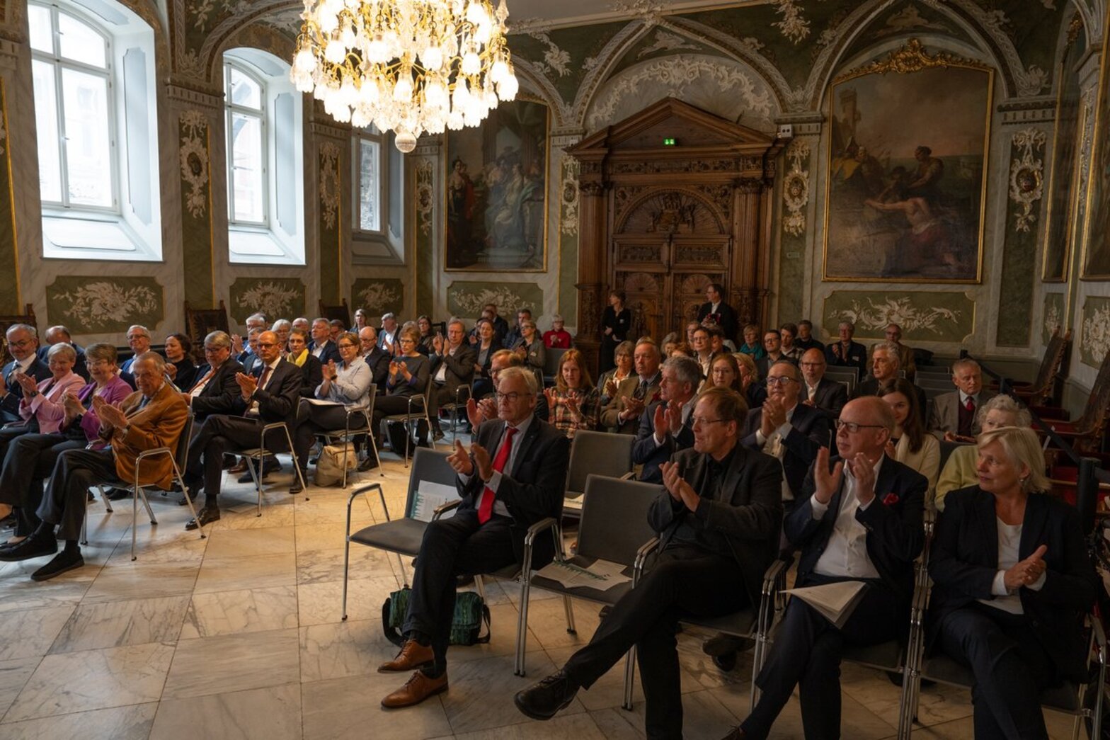 Eine Reihe von Menschen sitzt in einem barocken festlichem Saal, zwei Kronleuchter hängen von der Decke