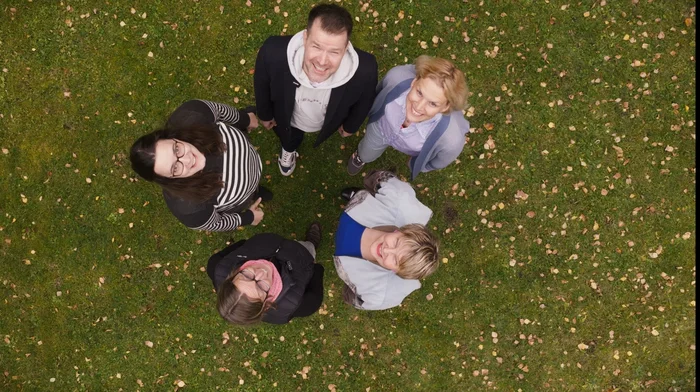 Eine Gruppe Menschen steht im Kreis und schaut nach oben und wird von einer Drohne fotografiert. 