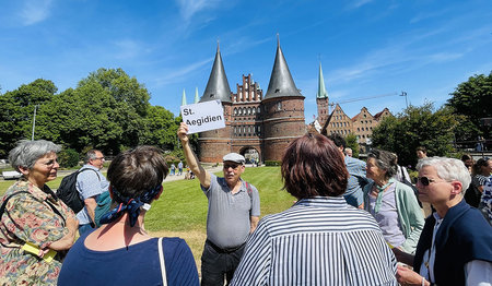 Herr Thurau steht vor der Graswiese vom Holstentor und hält ein Schild auf dem St. Aegidien steht hoch. Im Vordergrund ist eine Reihe von Menschen versammelt. Der Himmel ist blau mit leichten Schleierwolken