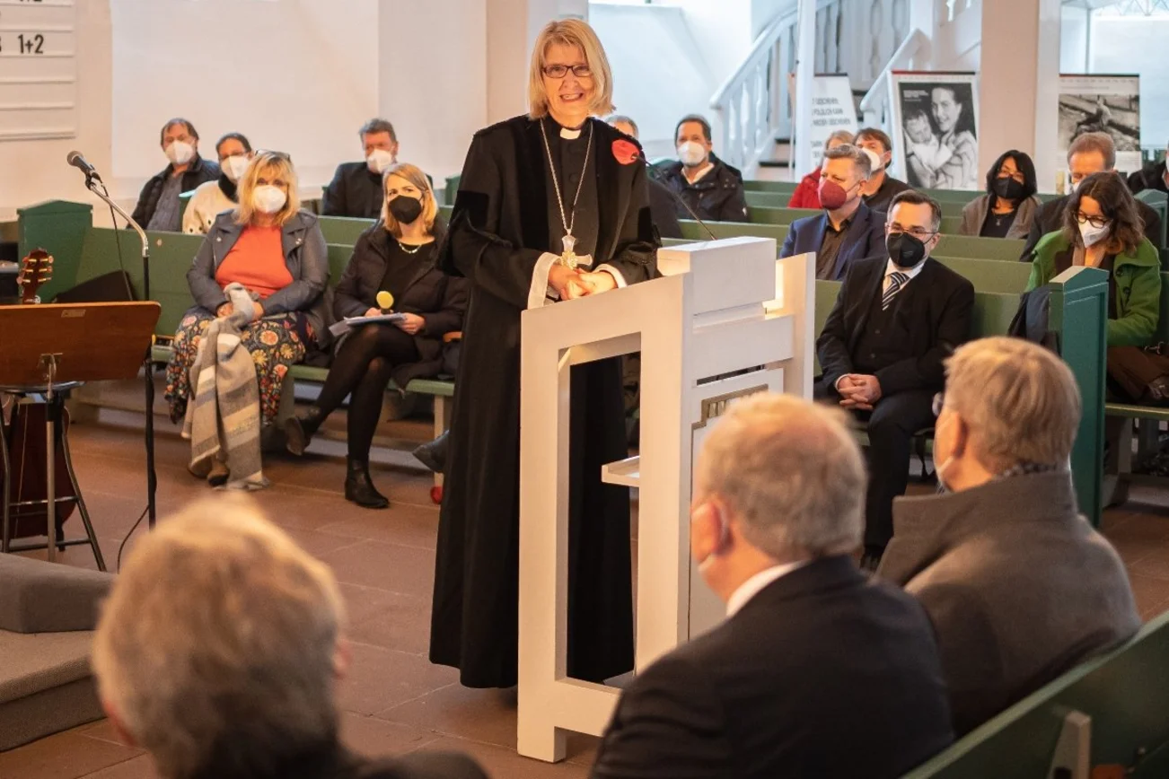 Pröpstin Frauke Eiben spricht am Rednerpult in ein Mikrofon. Menschen sitzen in Bänken und sind von hinten zu sehen.