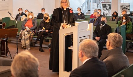 Pröpstin Frauke Eiben spricht am Rednerpult in ein Mikrofon. Menschen sitzen in Bänken und sind von hinten zu sehen.