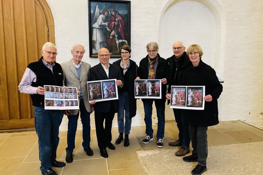 Das restaurierte Gemälde hängt an der Wand im Dom zu Lübeck, davor steht eine Gruppe Menschen - die Beteliligten. Sie halten Ausdrucke der Arbeitsschritte in den Händen und lächeln in die Kamera.  - Copyright: Steffi Niemann