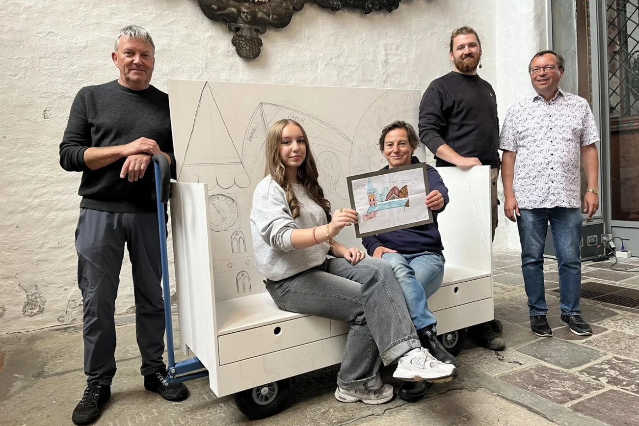 Fünf menschen sitzen auf und stehen um die noch unbemalte Kirchenbank herum und lächeln in die Kamera