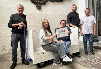 Fünf menschen sitzen auf und stehen um die noch unbemalte Kirchenbank herum und lächeln in die Kamera - Copyright: Katharina Micheel