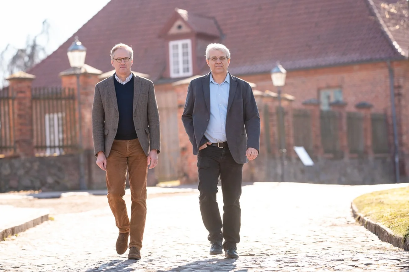 Zwei Männer gehen nebeneinander auf einem Weg mit Kopfsteinpflaster. Im Hintergrund ein Backsteingebäude. 