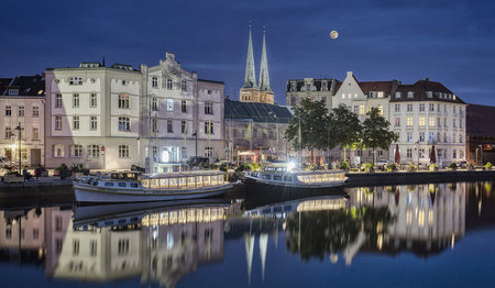 Der dunkle Abendhimmel bildet einen schönen Kontrast zu den beleuchteten Türmen des Doms im Hintergrund. Die schöne Häuserreihe an der Obertrabe wird von den Lichtern der Straße und Promenade leicht beleuchtet. Die Obertrave liegt ganz ruhig und spiegelt diese schönen Lichter. Auf der Trave liegen zwei eingeschossige Rundfahrtschiffe am Kai