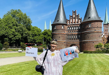 Frau Kramp hält in der linken Hand ein Schild auf welchem St. Marien steht, in der rechten das Logo vom Sieben-Türme-Projekt. Im Hintergrund ist das Holstentor und rechts daneben St. Petri zu sehen. Ein klarer blauer Himmel und Sonnenschein - Copyright: Steffi Niemann