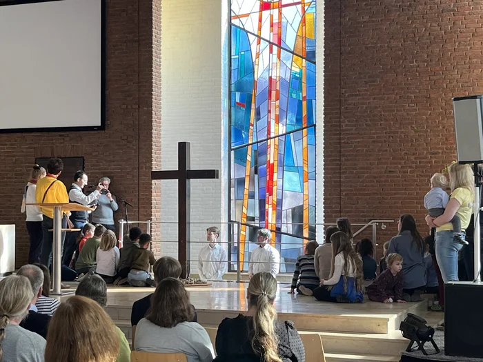 Mehrere Menschen in einer Kirche-2 Personen stehen hinter einem Kreuz in einem Taufbecken, andere stehen darum herum und einige auf Stühlen vor ihnen
