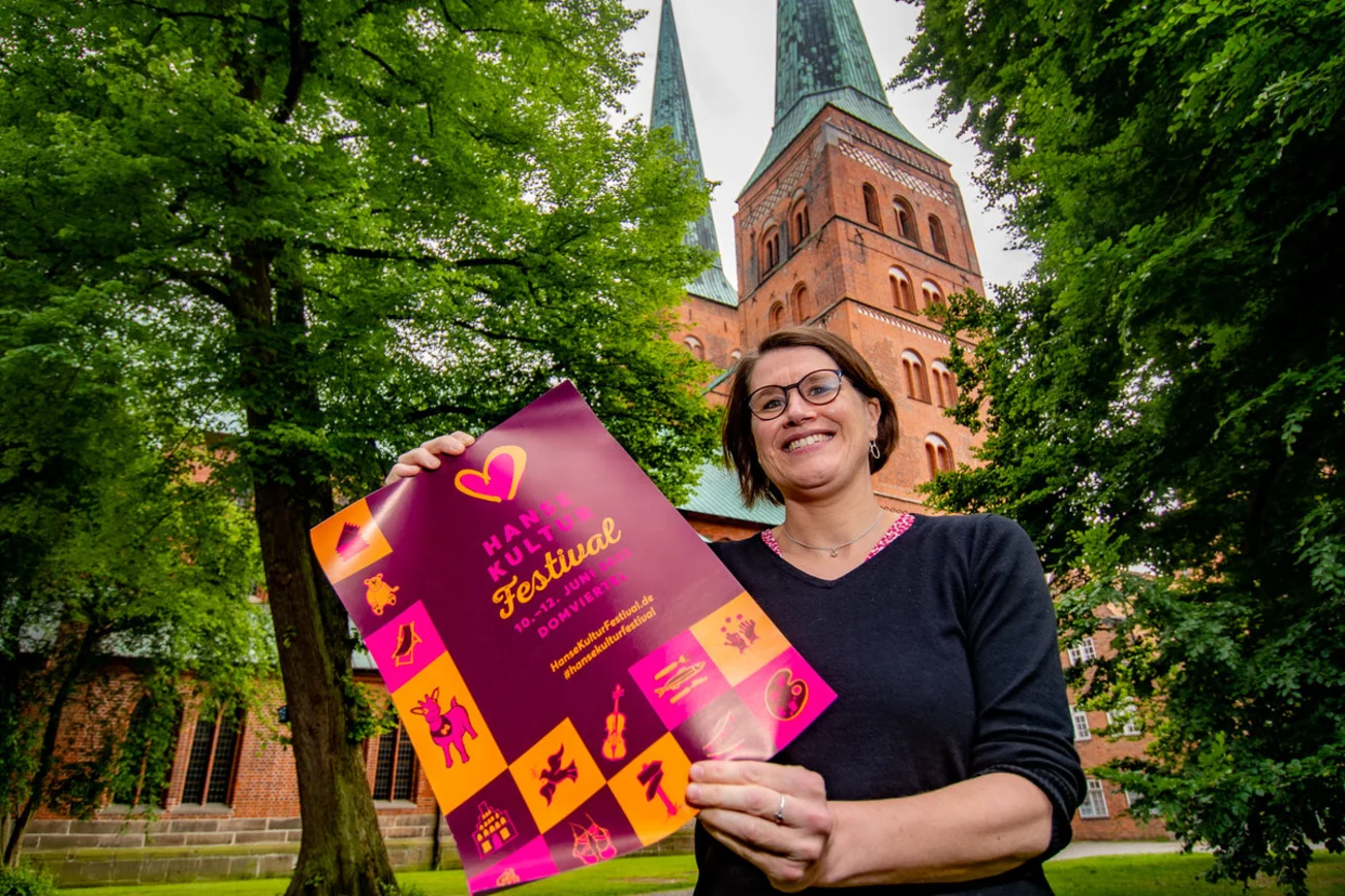 Eine Frau hält ein Plakat vor einer Kirche in die Kamera und lächelt. 